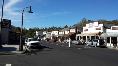 A quaint street scene with shops and cars under a clear blue sky, featuring a mix of storefronts and a pedestrian.