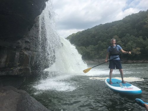A person stands on a paddleboard near a waterfall, surrounded by lush greenery and rocky cliffs.