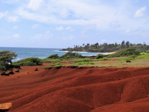 A scenic view of red soil hills leading to a coastline with waves and lush greenery under a blue sky.