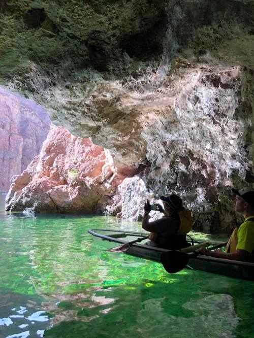 A person in a kayak takes a photo inside a rocky cave with green water and illuminated rock formations.