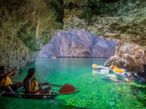 Two kayakers explore a scenic cave with turquoise water and rocky cliffs in the background. Sunlight filters through.