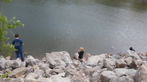 Two people fishing by a rocky riverbank, with one sitting on the rocks and the other standing nearby.