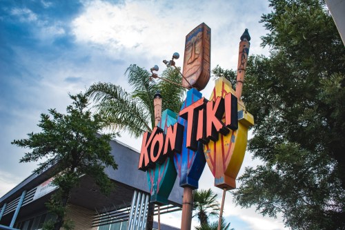 Colorful "Kon Tiki" sign with tiki motifs and palm trees against a cloudy sky.