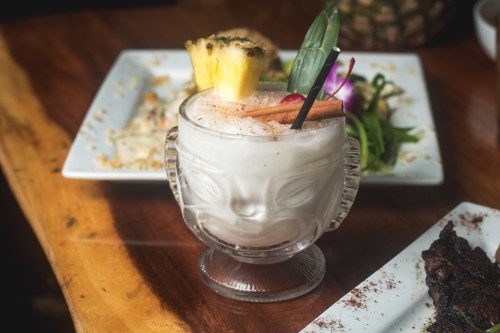 A tropical cocktail in a decorative glass, garnished with pineapple, a cherry, and a cinnamon stick, on a wooden table.