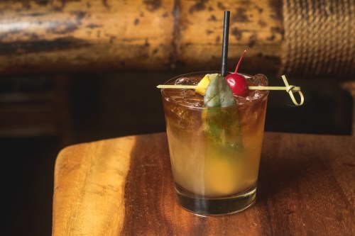 A refreshing cocktail in a glass with ice, garnished with a cherry, lemon, and a green leaf, on a wooden table.