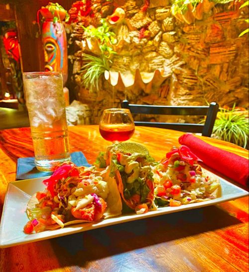 A plate of three colorful tacos with toppings, a glass of iced drink, and a small glass of brown liquor on a wooden table.