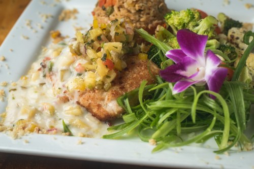 A plated dish featuring grilled fish topped with salsa, surrounded by vegetables and garnished with an orchid flower.