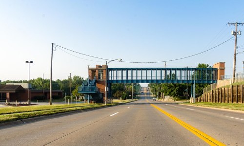 A clear view of a road leading to a pedestrian overpass, with buildings and trees in the background under a blue sky.