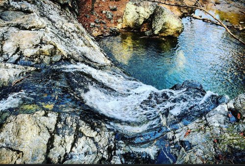 A small waterfall cascades over rocky terrain into a serene pool, surrounded by natural scenery.