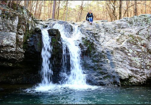 A person sits on a rock near a waterfall cascading into a serene pool, surrounded by trees and rocky terrain.