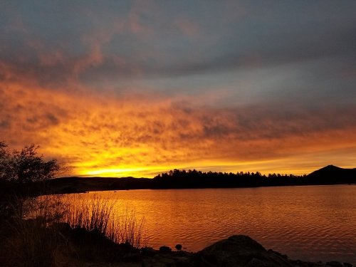 A vibrant sunset over a calm lake, with silhouettes of trees and hills against a colorful sky.