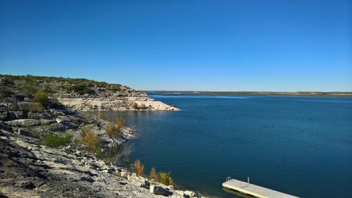 A serene lake view with rocky shores and clear blue skies, featuring a small dock extending into the water.