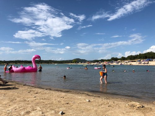 A sunny beach scene with people swimming, a large pink flamingo float, and a sandy shore under a blue sky.
