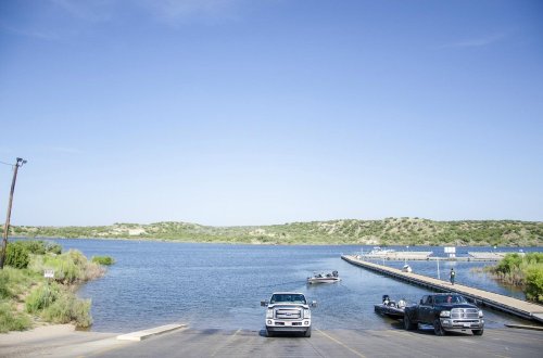 A boat ramp by a calm lake, with two trucks parked and a boat being launched under a clear blue sky.