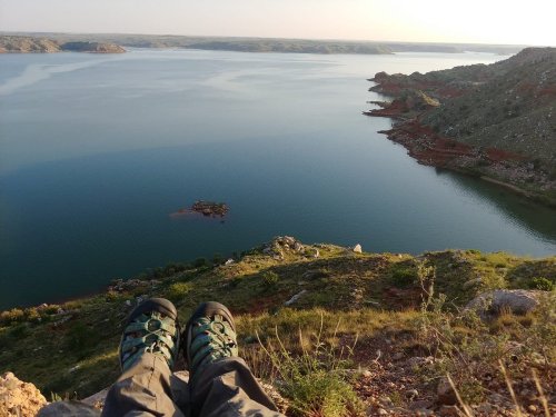Person's feet in hiking boots resting on a rocky ledge, overlooking a calm lake surrounded by hills and greenery.