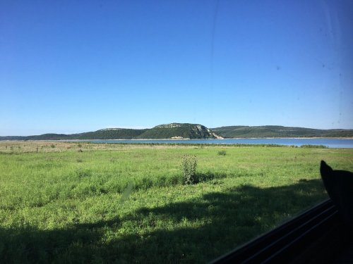 A serene landscape featuring a green field, a calm lake, and a distant hill under a clear blue sky.