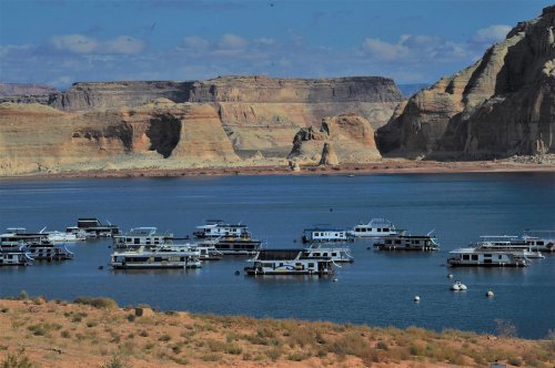 A scenic view of houseboats anchored in a calm lake, surrounded by rocky cliffs and blue skies.