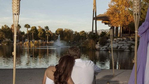A couple sits together by a serene lake, surrounded by palm trees and autumn foliage, enjoying a peaceful moment.