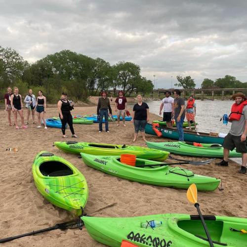 A group of people stands on a sandy beach with green kayaks and a river in the background, under a cloudy sky.