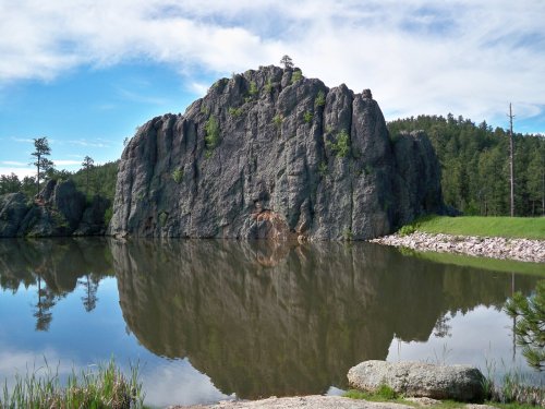 A large rock formation reflected in a calm lake, surrounded by green trees and a clear blue sky.