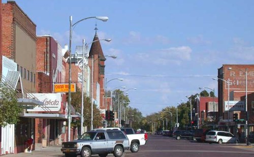 A small town street lined with brick buildings, shops, and parked cars under a clear blue sky.