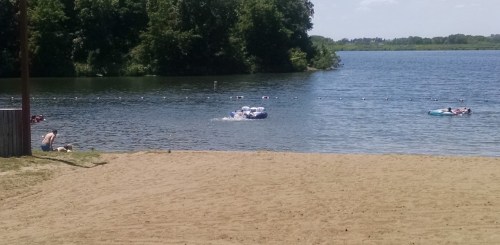 A sandy beach by a lake with people swimming and floating on rafts in the water, surrounded by trees.