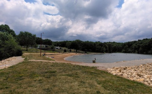 A scenic view of a lake surrounded by trees, with sandy areas and people enjoying the water under a cloudy sky.