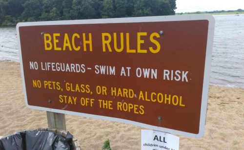 Sign at a beach listing rules: no lifeguards, swim at own risk, no pets or glass, and stay off the ropes.