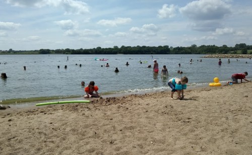 A sandy beach with children playing in the water and on the shore, under a sunny sky with scattered clouds.