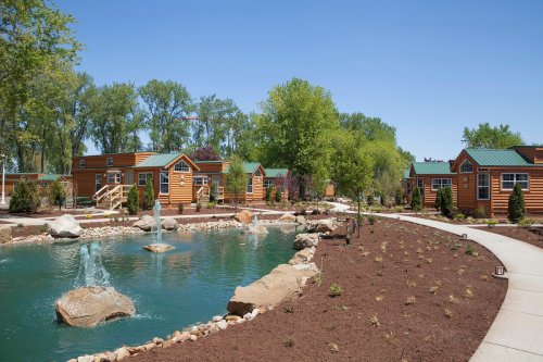 A serene landscape featuring wooden cabins by a pond, surrounded by greenery and a clear blue sky.