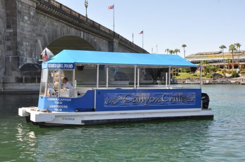 A tour boat named "Canyon Cruiser" with a blue canopy, docked near a bridge and palm trees by the water.