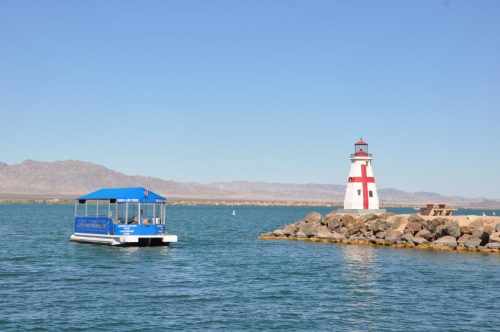 A boat near a lighthouse on a calm lake, with mountains in the background under a clear blue sky.