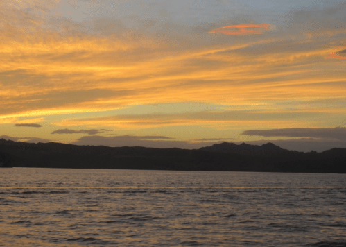 A serene sunset over calm waters, with vibrant orange and yellow hues illuminating the sky and silhouetting distant mountains.