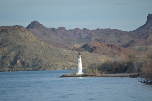 A white lighthouse stands on a small island in a lake, surrounded by mountains and a clear blue sky.