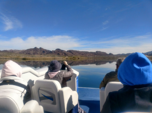 A group of people on a boat, facing a calm lake surrounded by mountains under a clear blue sky.