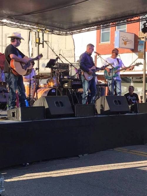A live band performs on stage with instruments, under a canopy, in a lively outdoor setting.
