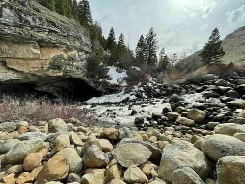 A rocky riverbank with boulders, flowing water, and a cave entrance surrounded by trees and snow.