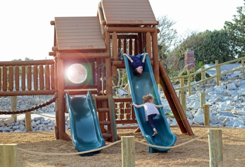 A child slides down a green slide while an adult slides down from above at a wooden playground. Sunlight shines in the background.