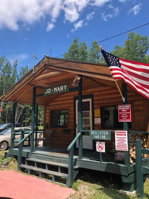 A rustic log cabin with a sign reading "Jo-Mary," an American flag, and a notice about registration and fireworks.