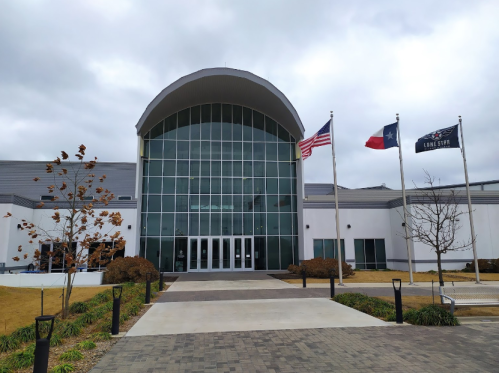 Modern building entrance with large glass facade, flanked by flags, and landscaped pathway leading up to it.