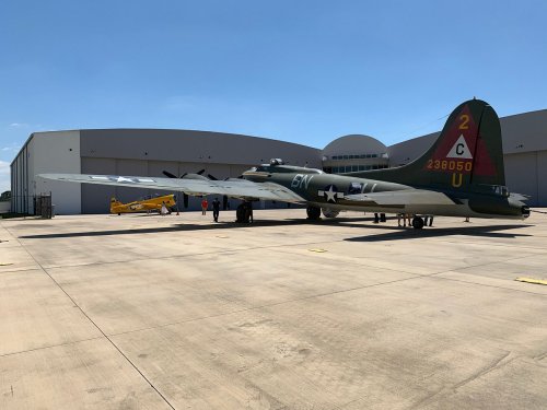 A vintage B-17 bomber and a yellow biplane parked outside a hangar under a clear blue sky.