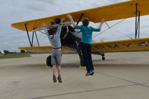 Two children jump in front of a vintage biplane on a cloudy day, arms raised in excitement.