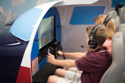 Two children wearing headsets sit in a flight simulator, focused on the screen in front of them.