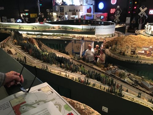 A detailed model train layout with trees, tracks, and people observing the scene in a museum setting.