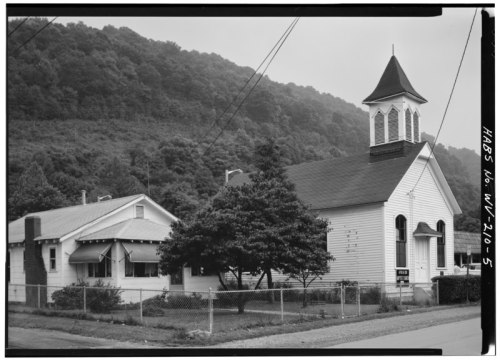 Historic church and adjacent building surrounded by trees, with a mountainous backdrop and a fence in the foreground.