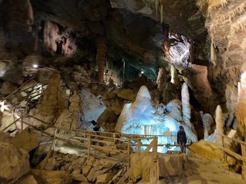 A dimly lit cave with rock formations and a person standing near a blue-lit stalagmite. Wooden walkways are visible.