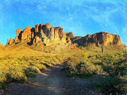 A scenic desert landscape featuring rugged mountains under a clear blue sky, with a dirt path leading through green vegetation.