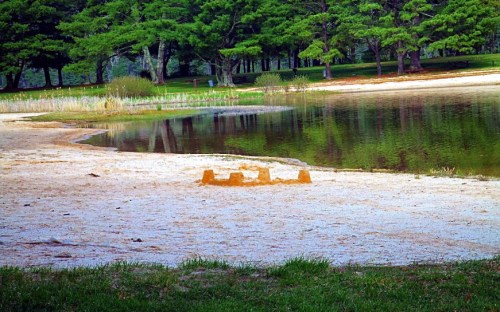 A sandy beach by a calm lake, featuring a small sandcastle and lush green trees in the background.