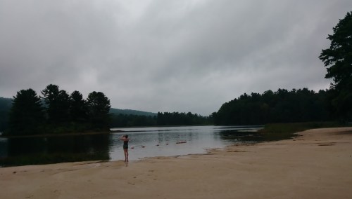 A person stands on a sandy shore by a calm lake, surrounded by trees under a cloudy sky.