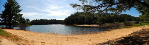 A serene lake surrounded by trees, with a sandy beach and clear blue sky, creating a peaceful natural landscape.